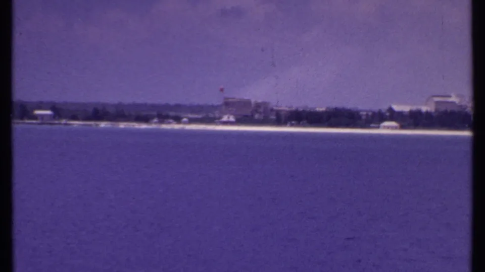 1969 Bahamas 8mm frame with blue water in front, distant shore structures, trees, and a pale sandy edge beneath a hazy sky.