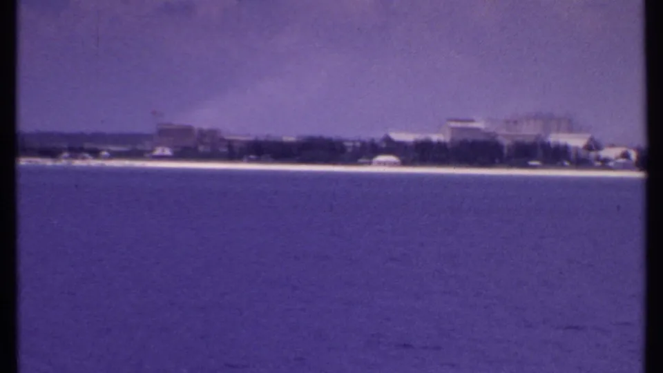 1969 Bahamas coastal frame showing larger distant waterfront structures, a narrow beach, and calm water with visible 8mm grain and color shift.
