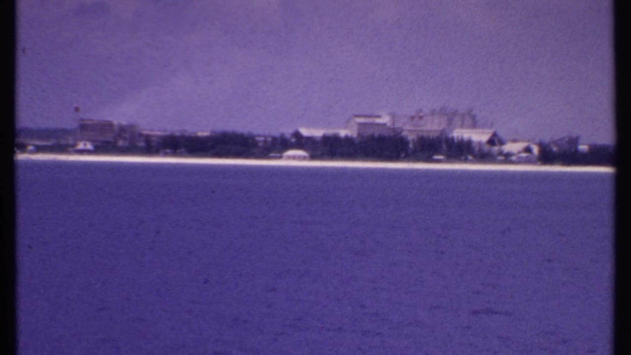 1969 8mm view from the water toward a quiet Bahamas shoreline with low buildings, trees, sandy coast, and hazy sky.