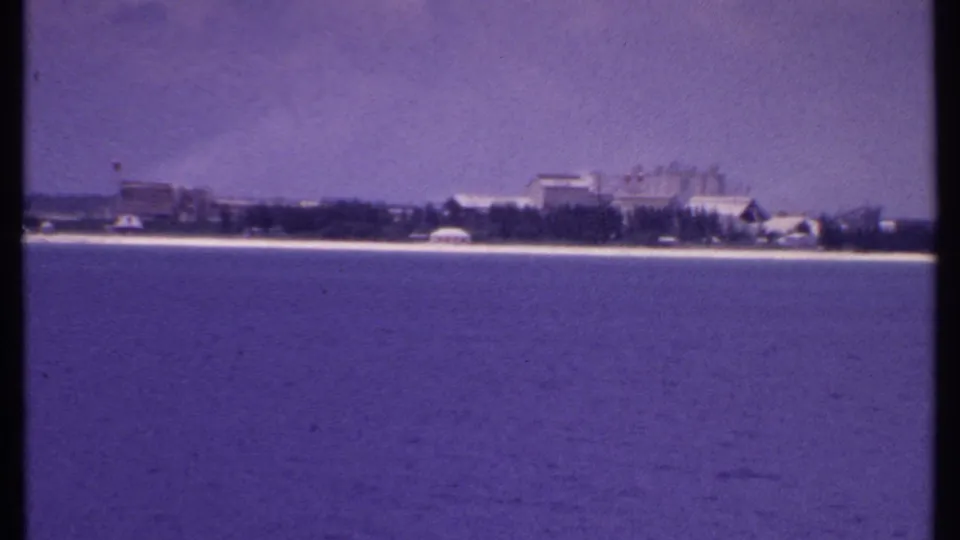 1969 8mm view from the water toward a quiet Bahamas shoreline with low buildings, trees, sandy coast, and hazy sky.