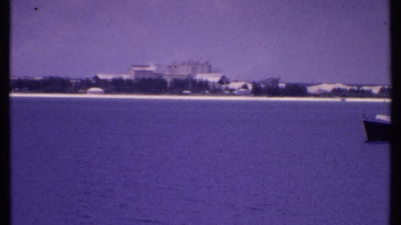 1969 Bahamas shoreline seen across open water, with distant buildings and a partial dark boat entering at the right edge.