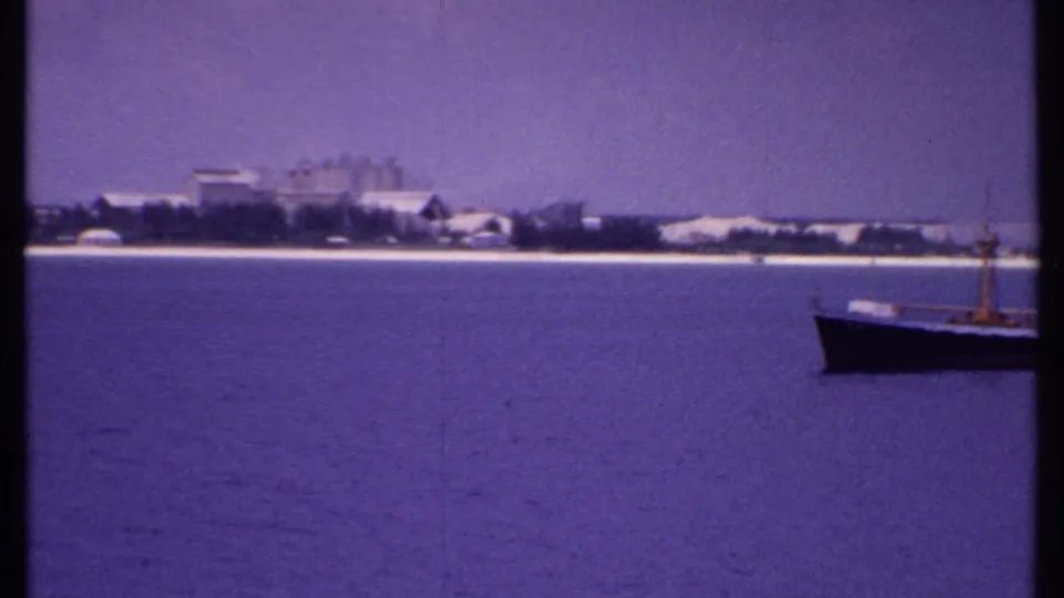 Small boat moves across calm water before a distant 1969 Bahamas coastline with pale beach, trees, and scattered buildings.