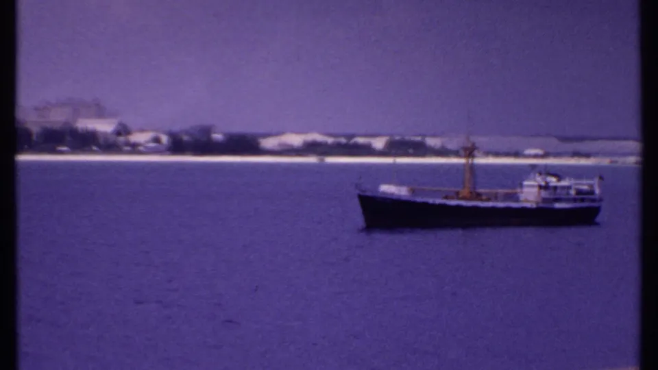 1969 8mm frame of a small boat on blue water, set against a quiet Bahamas shoreline with sparse buildings and trees.