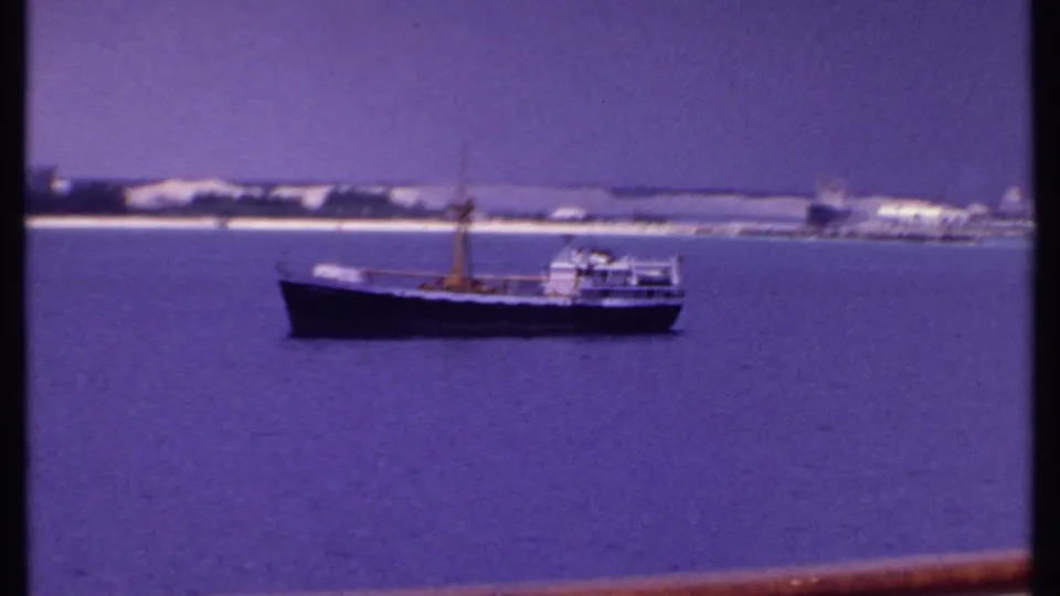 Small boat seen from a cruise ship deck in the Bahamas, with purple-blue water, distant shore, and a red-brown rail crossing the frame.