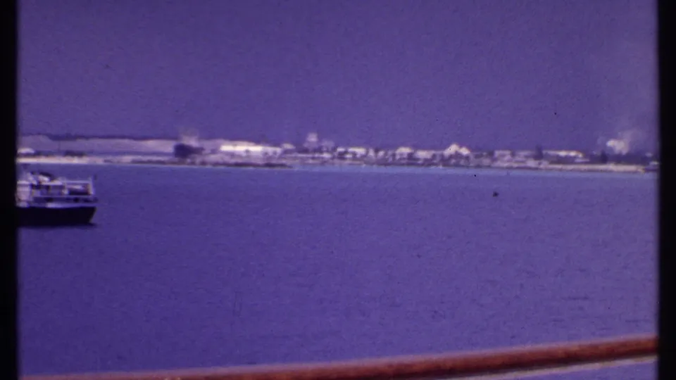 The camera pans past the boat, leaving it partly cropped while the Bahamian shoreline and ship rail dominate the view.