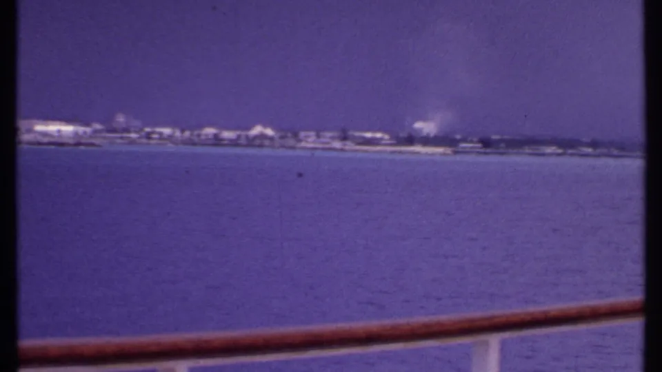 Cruise-deck view across calm Bahamian water toward a low shoreline, with a faint smoke plume rising under a hazy sky.