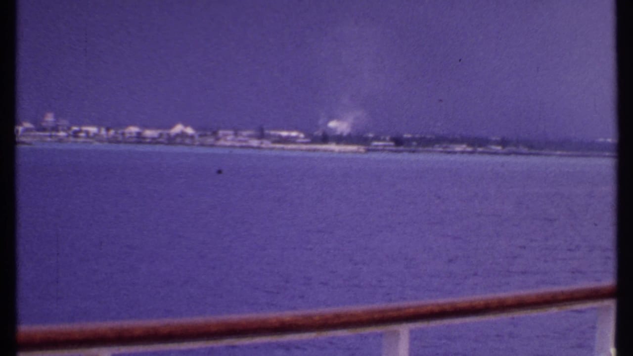 1969 Bahamas boat view across calm water toward a low, hazy coastline, framed by a wooden ship rail.