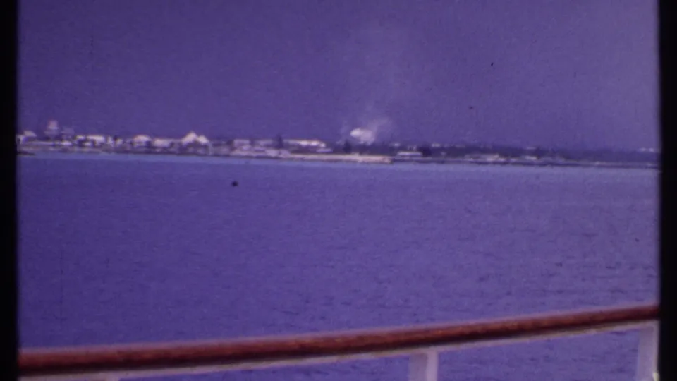 1969 Bahamas boat view across calm water toward a low, hazy coastline, framed by a wooden ship rail.
