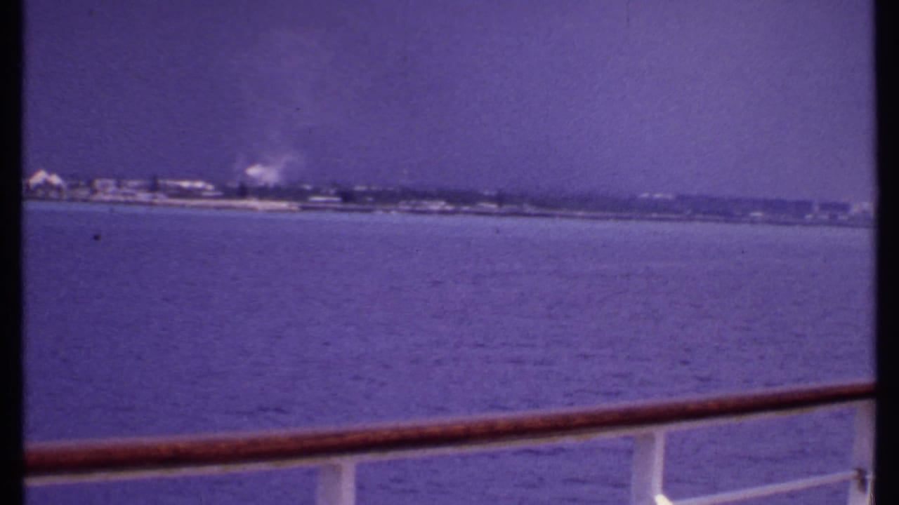 1969 Bahamas home-movie frame showing calm water and indistinct coastal buildings beyond a wooden ship rail.