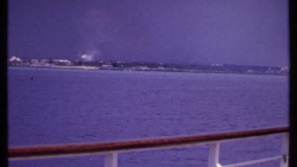 1969 Bahamas home-movie frame showing calm water and indistinct coastal buildings beyond a wooden ship rail.
