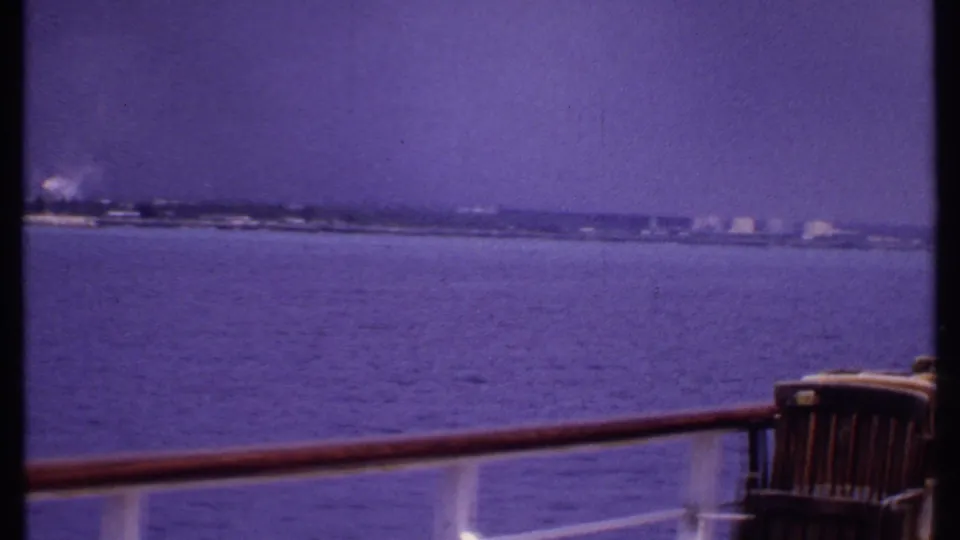 1969 Bahamas boat-deck view with wooden chairs, a rail, calm water, and a faint coastline in the distance.