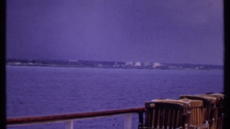 1969 Bahamas cruise-deck view with empty wooden chairs, rail, calm water, and a low distant coastline.