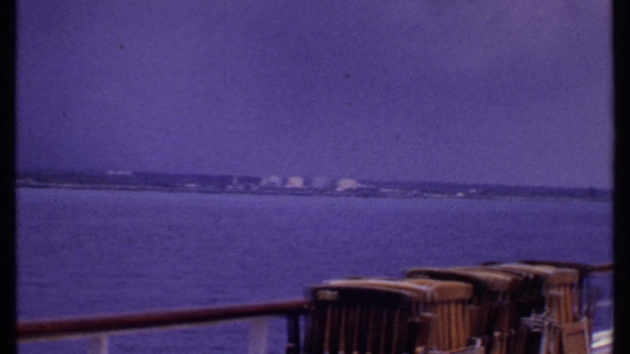 Wide sea view from a ship deck with distant shoreline and wooden deck chairs in the foreground.