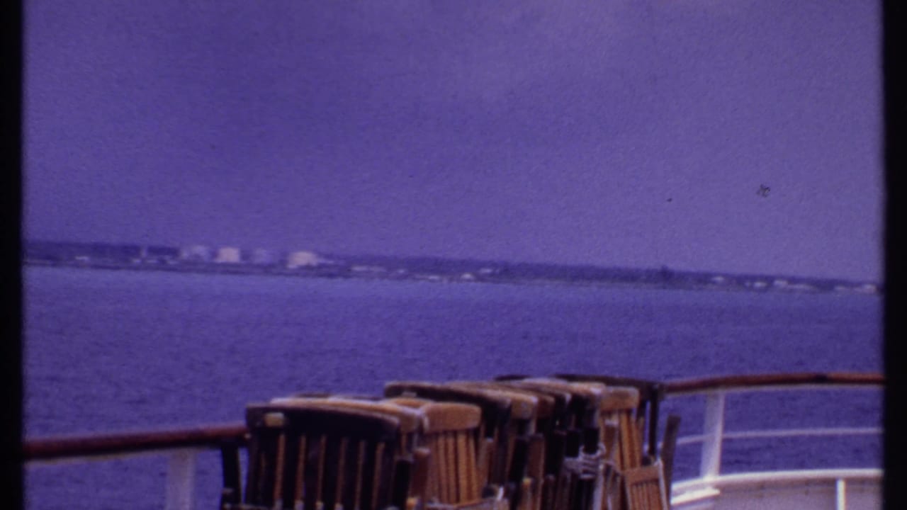 Ship deck view across open water toward a low shoreline with deck chairs and railing in front.