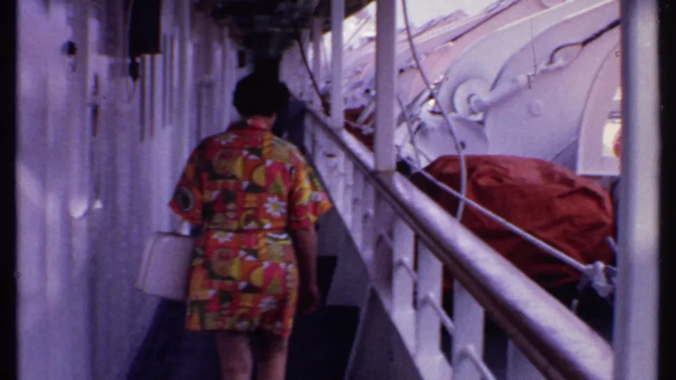 A woman in a bright patterned late-1960s dress walks away along a Bahamas cruise ship corridor, carrying a white purse.