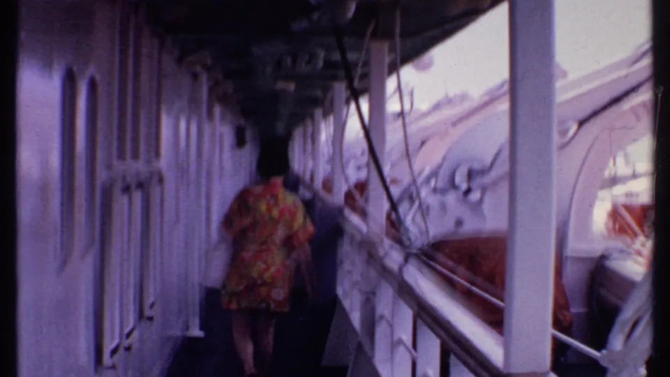 A passenger in a colorful dress walks through a shaded exterior deck passage with white ship fittings, railings, and covered boat gear visible.