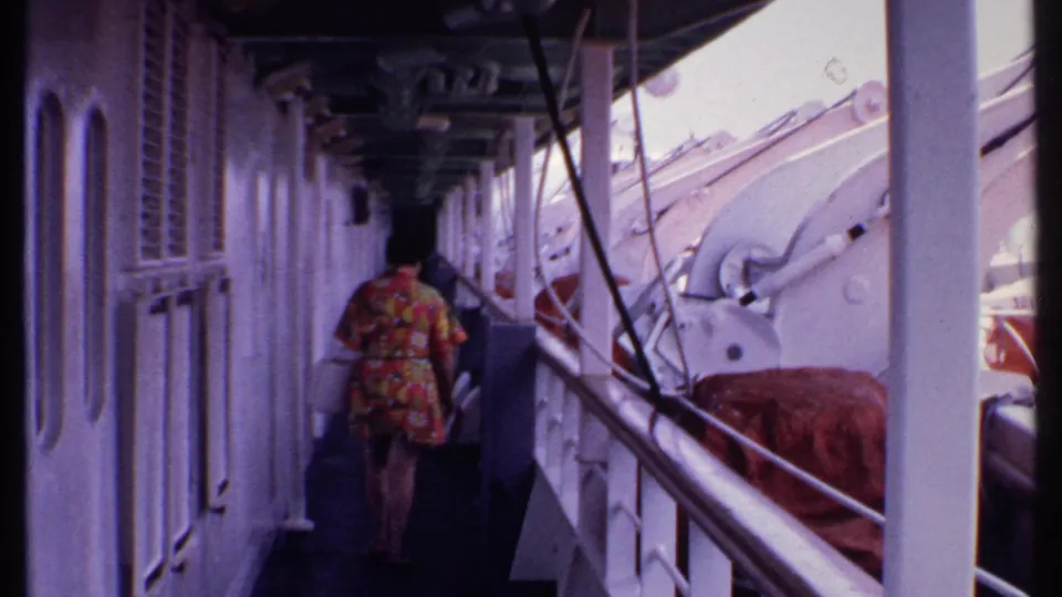 1969 Bahamas home-movie frame of a woman in a floral dress walking along a ship corridor beside railings and lifeboat equipment.