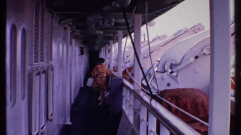 1969 Bahamas 8mm frame showing a person in a floral dress walking along a narrow covered ship deck with lifeboats nearby.