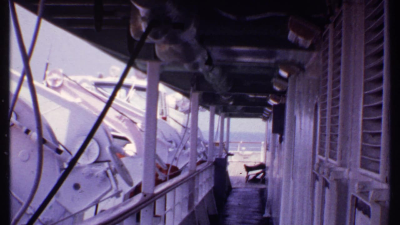 Covered ship passage with white lifeboat equipment, railings, windows, and sea at the far end.