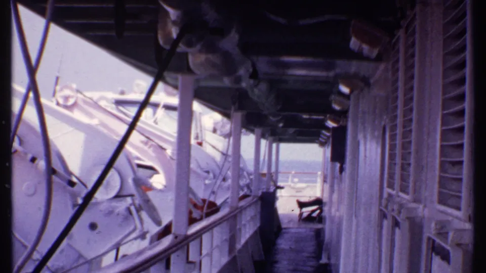 1969 Bahamas shipboard view down a covered deck passage with white lifeboats, railings, shuttered windows, and ocean beyond.