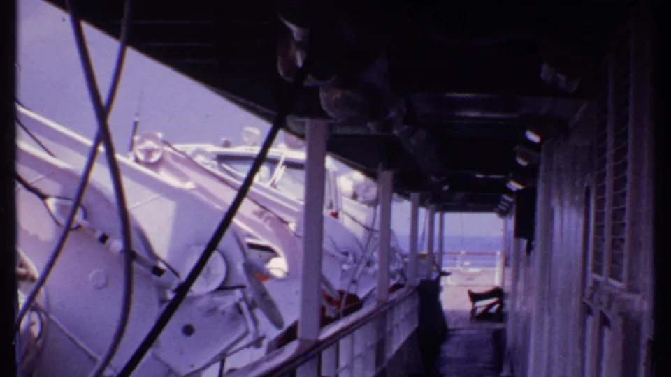 1969 Bahamas 8mm view of a ship deck corridor, with lifeboats, railings, overhead piping, and shuttered cabin windows.