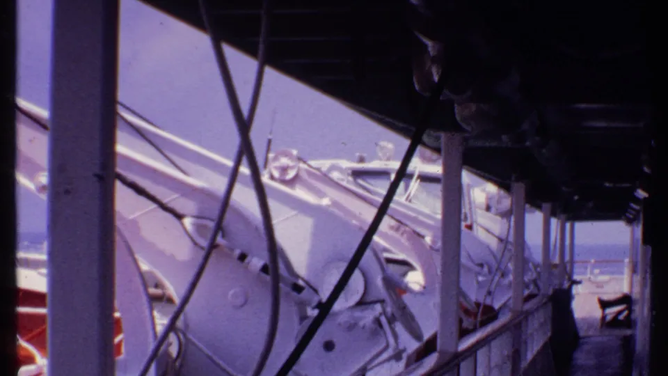 1969 Bahamas 8mm frame showing white boat equipment and railings from a shaded side passage facing the water.