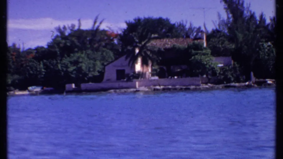 1969 Bahamas waterfront frame with blue water, dense green trees, and a small tiled-roof building along the shore.