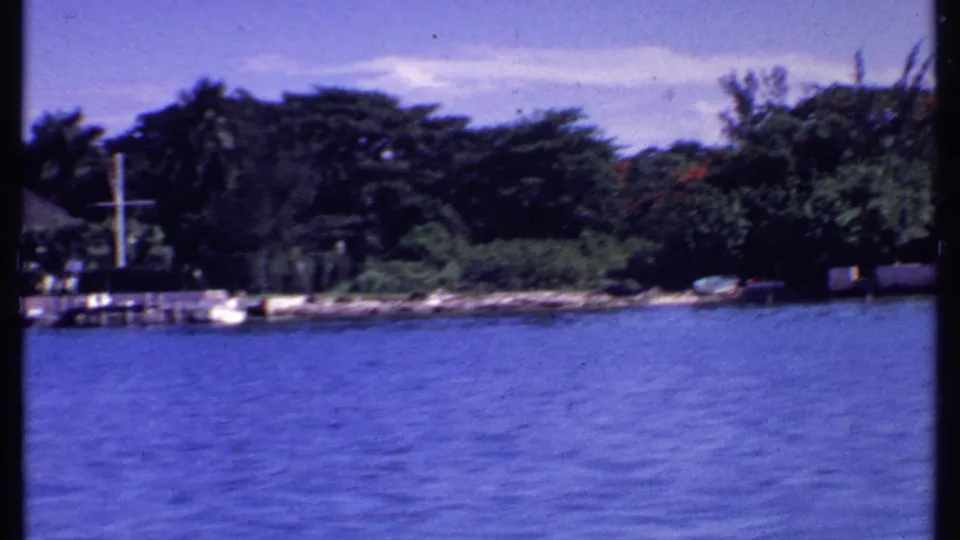 1969 Bahamas 8mm frame showing blue water, a tree-lined shore, a dock, and small boats near the waterfront.