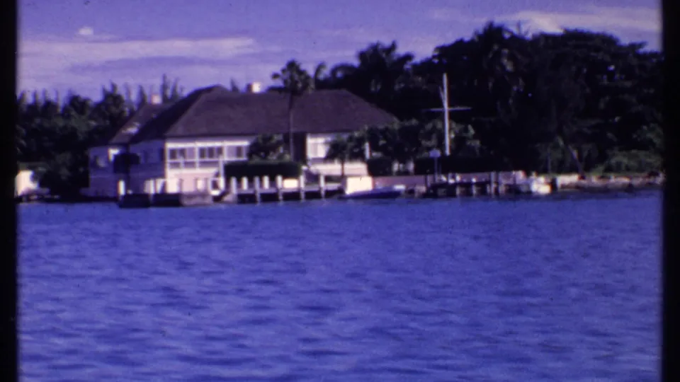 1969 Bahamas 8mm frame centered on a white waterfront house, dock, palms, and rippled water.