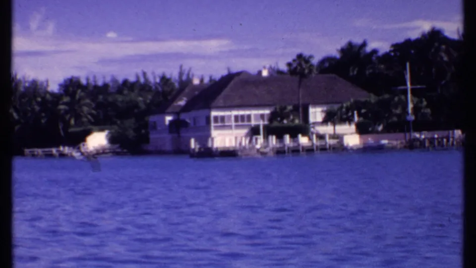 1969 Bahamas 8mm frame with a waterfront residence, dock, palms, and saturated blue water.