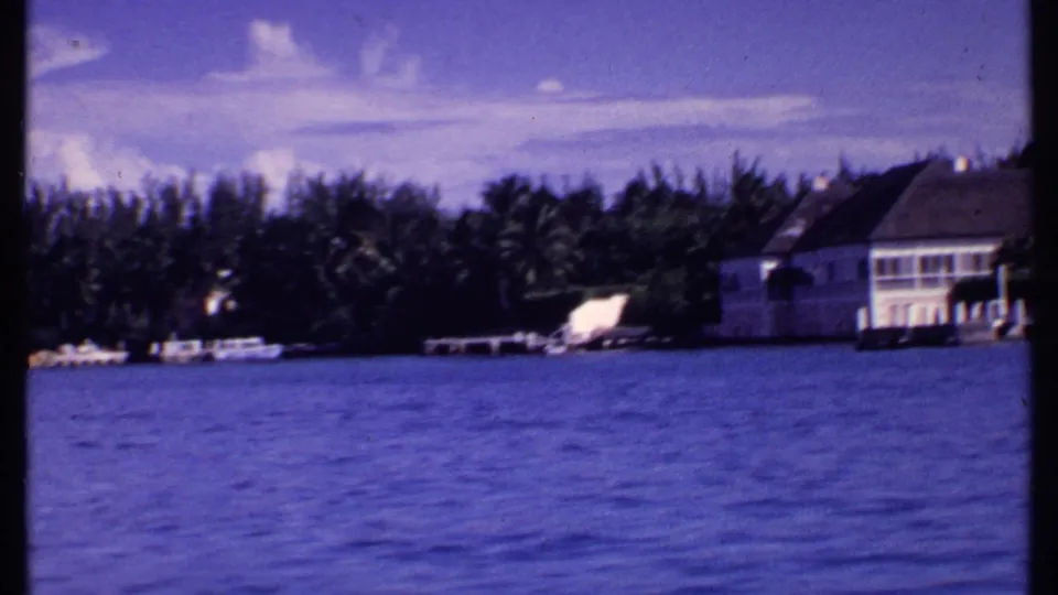 1969 Bahamas 8mm frame showing a partial waterfront house, dock, palms, and open blue water.