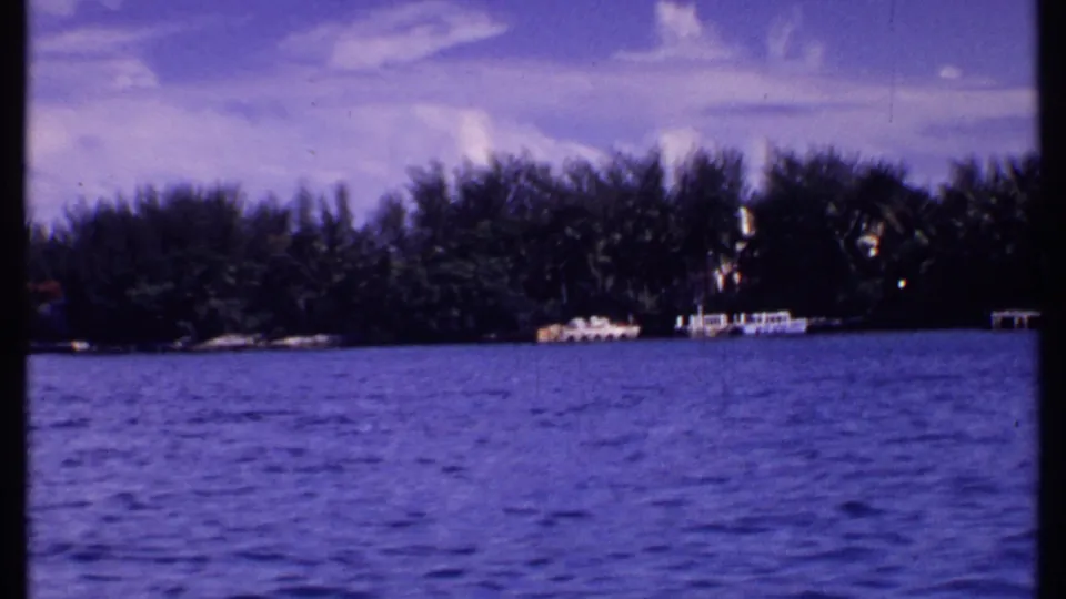 1969 Bahamas 8mm frame showing a broad water surface, distant shoreline trees, and small dock or boat forms in bright daylight.
