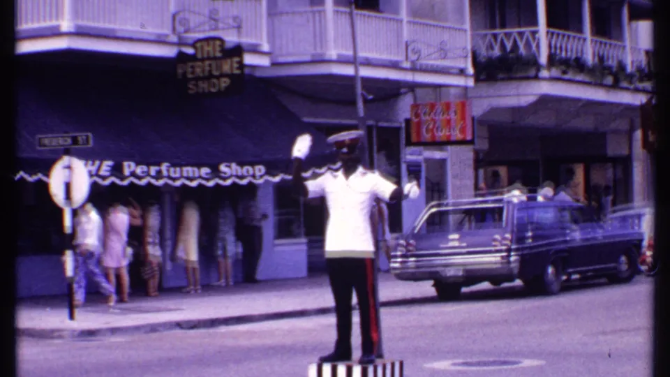 1969 Bahamas street corner with a white-shirted police officer directing traffic near The Perfume Shop, pedestrians, and a station wagon.