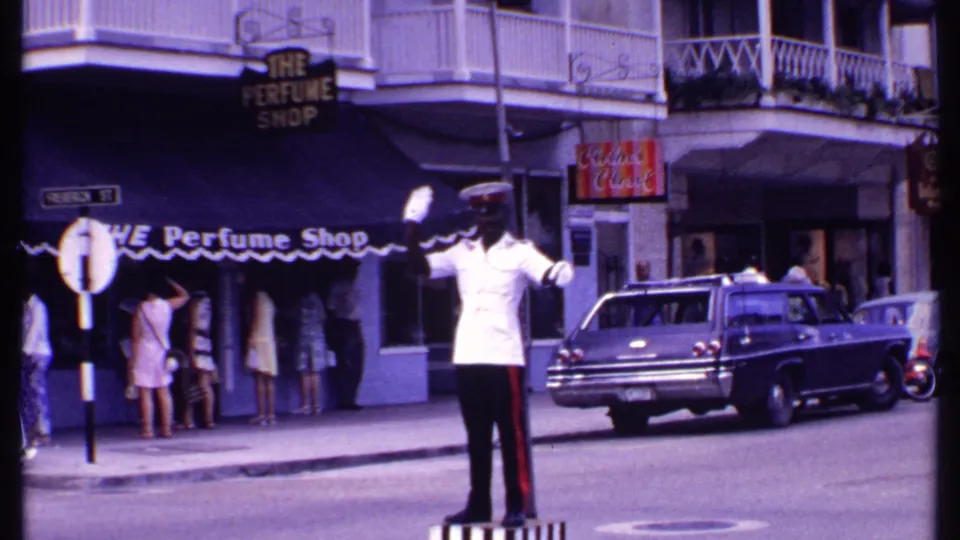 1969 Bahamas street view with a white-uniformed traffic officer signaling near shopfronts, pedestrians, and a parked station wagon.