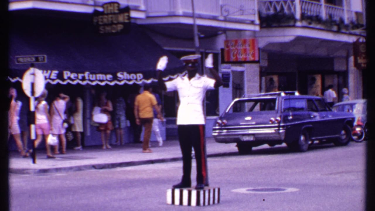 Officer raises both arms from a striped pedestal as pedestrians gather near shops and a station wagon waits at curbside.