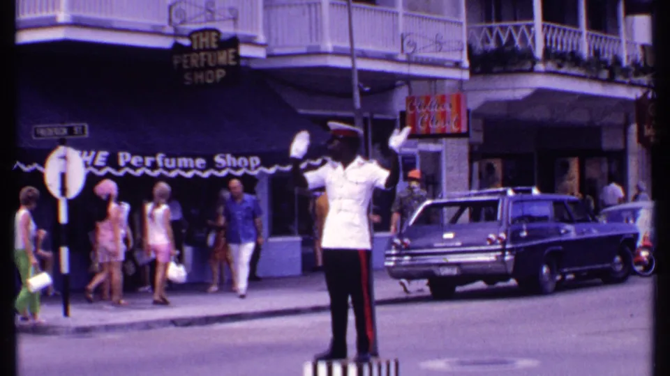 Street-level Bahamas footage shows the officer gesturing as pedestrians in 1960s dress pass The Perfume Shop and a parked wagon.