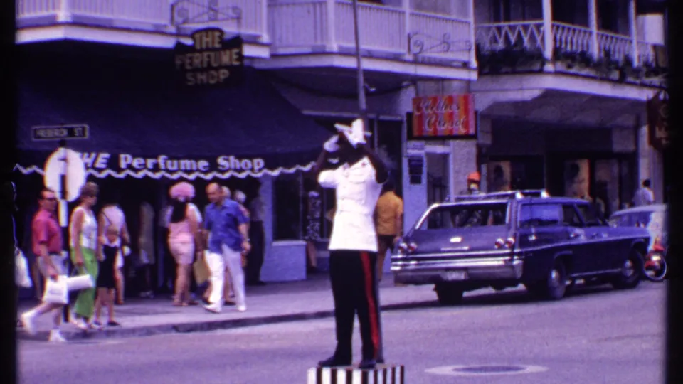 A white-uniformed Bahamas traffic officer directs movement near shop signs, pedestrians, balconies, and a 1960s station wagon.