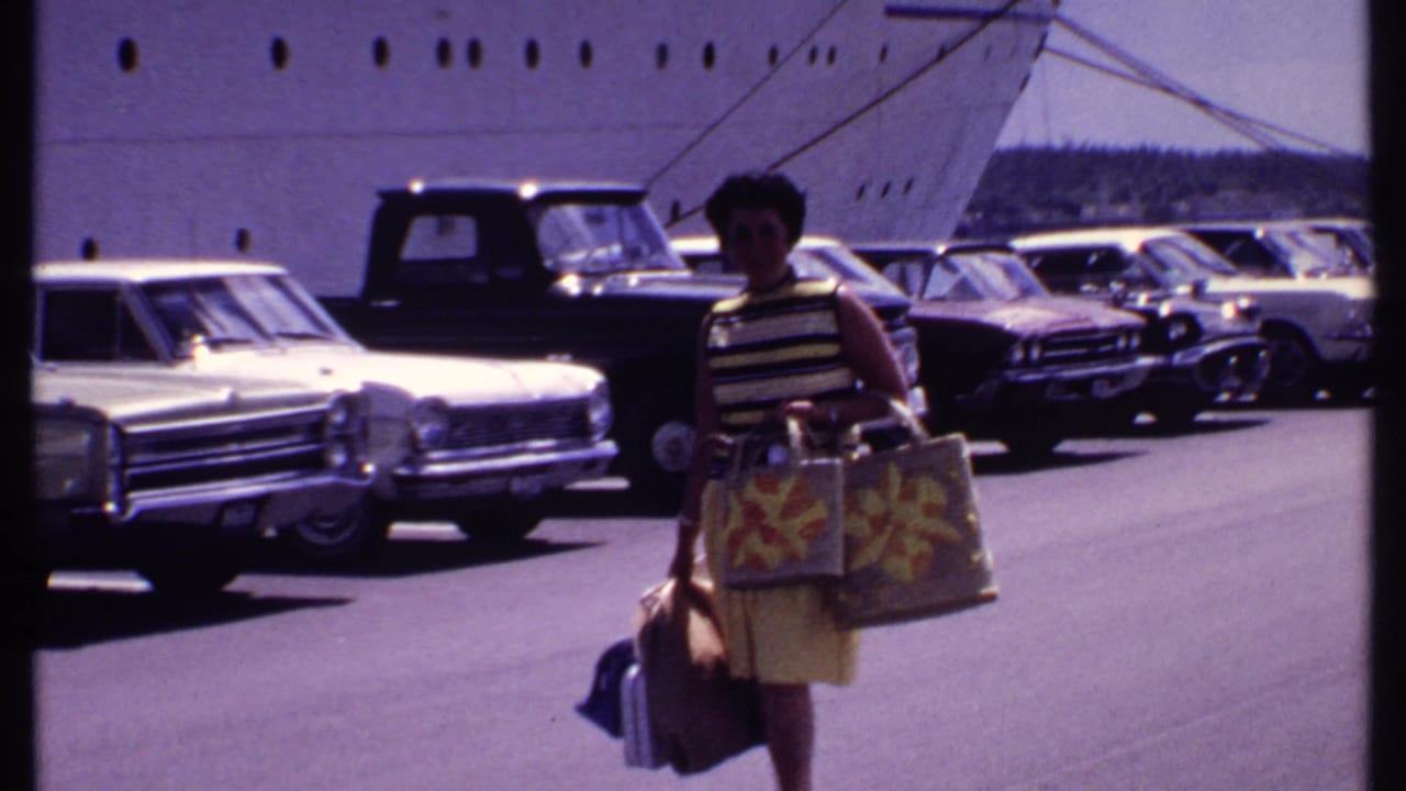 Woman carrying bags in a sunny parking lot beside a large cruise ship and classic cars.