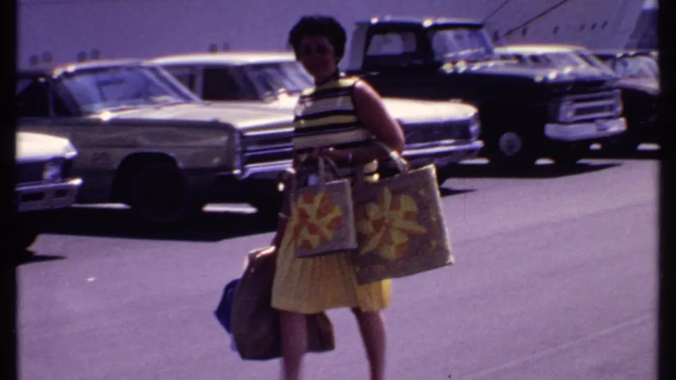 A woman carrying patterned bags and luggage appears near late-1960s cars with a cruise ship visible behind her in the Bahamas.