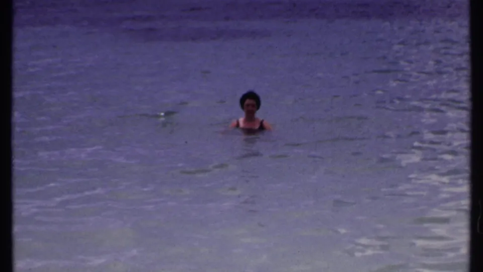Adult woman in a dark bathing suit faces the camera while bobbing in calm Bahamas ocean water in 1969 home-movie footage.