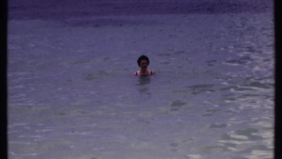The woman remains chest-deep in calm Bahamas water, centered in a soft 8mm frame with no visible shoreline detail.