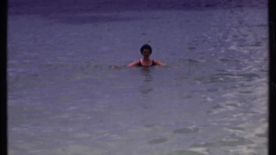 An adult woman in a dark bathing suit spreads her arms slightly while floating in calm 1969 Bahamas ocean footage.