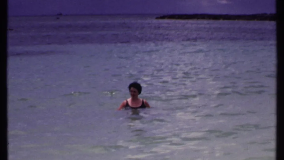 The swimmer bobs in blue-green Bahamas water with a low distant shoreline visible near the horizon in the 1969 home-movie frame.