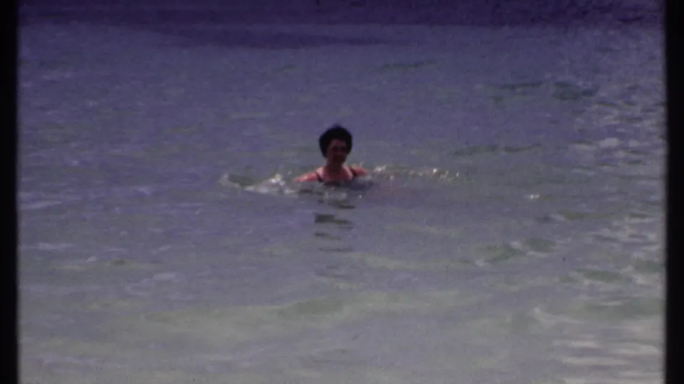 Woman swims toward or across the camera’s view in calm Bahamas water, with visible 8mm grain and color shift.