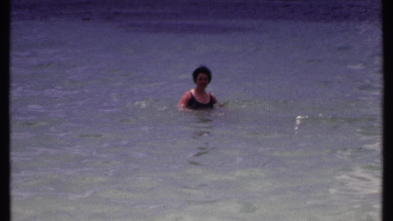 Adult woman in a dark swimsuit faces the camera while swimming in shallow Bahamas water in 1969 home-movie footage.