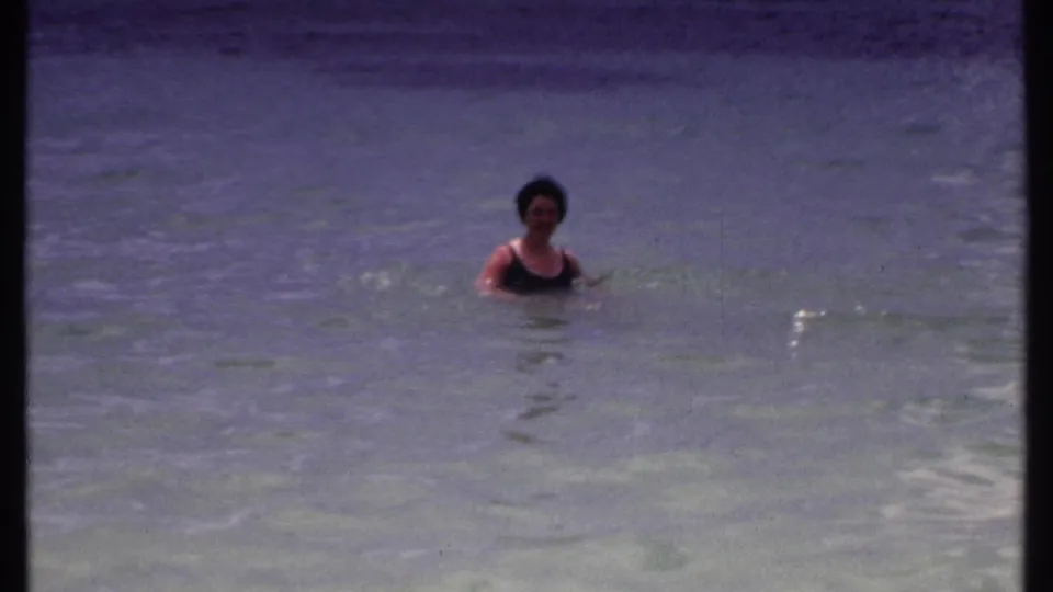 Adult woman in a dark swimsuit faces the camera while swimming in shallow Bahamas water in 1969 home-movie footage.