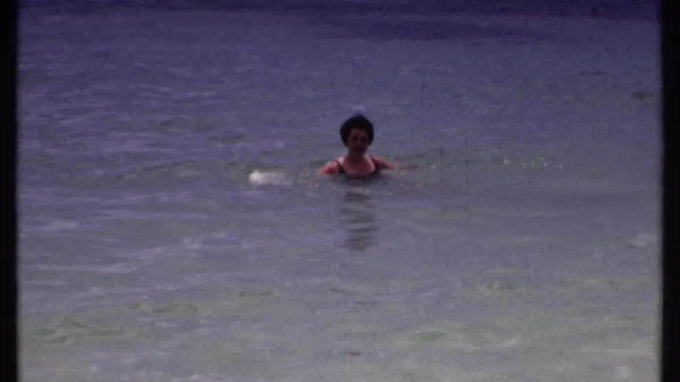 Adult woman swims with her head and shoulders above calm tropical water in the 1969 Bahamas clip.