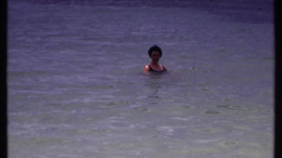 Adult woman remains upright in calm shallow water, framed by the purple-blue cast of 1969 8mm Bahamas footage.