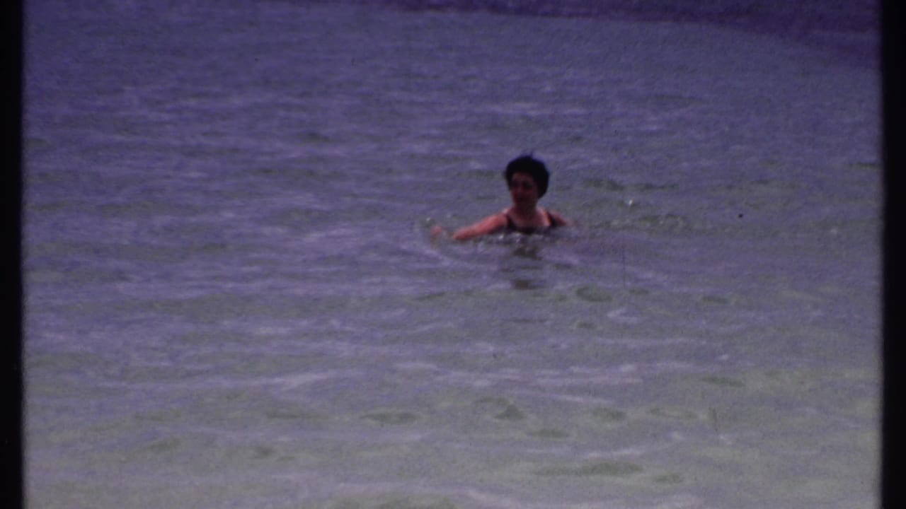 Adult woman in a dark swimsuit swimming in shallow blue-green water.