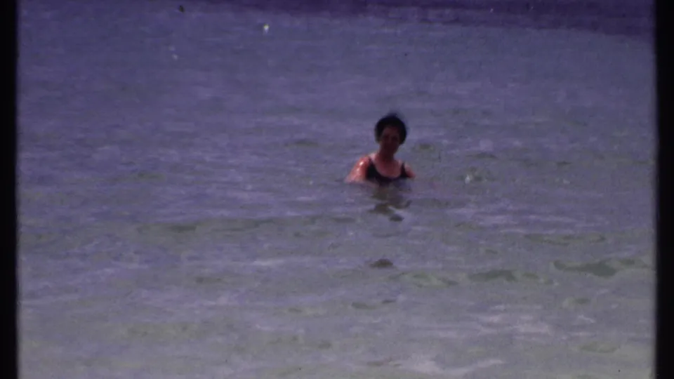 The swimmer remains chest-deep in calm Bahamas water, filmed from a distant 8mm home-movie viewpoint.